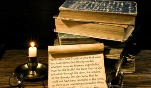 Old books and a scroll with writing on it and a quill next to a burning candle on a candleholder.